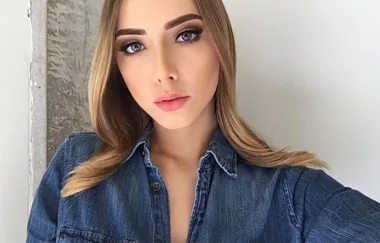 A person with long blonde hair wearing a denim shirt poses against a white and textured wall background.
