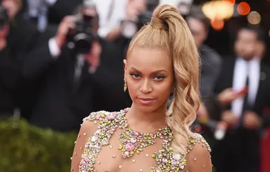 A woman with a high ponytail and embellished dress poses on a red carpet, photographers in the background.