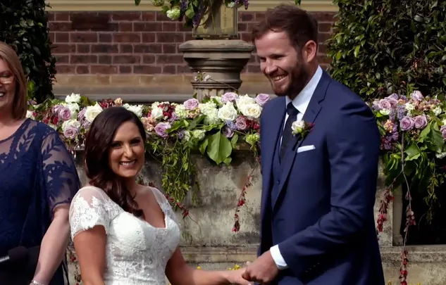 A couple smiling at their wedding ceremony, holding hands in front of floral arrangements.