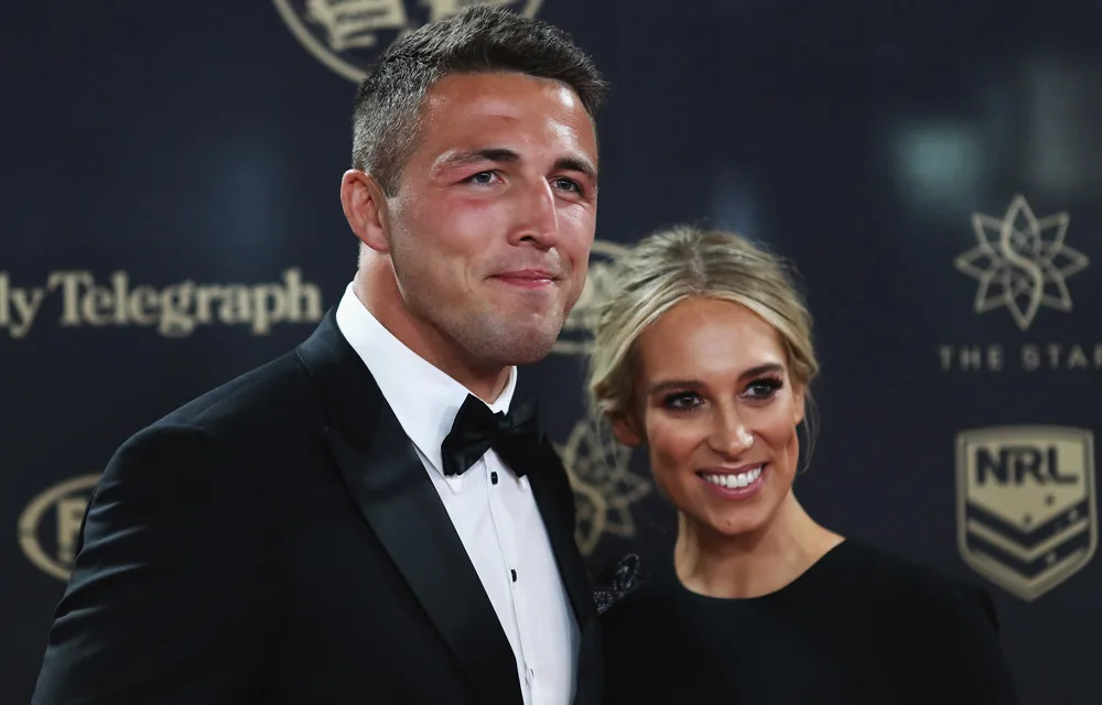 Couple in formal attire smiling at an event with a dark backdrop featuring logos.