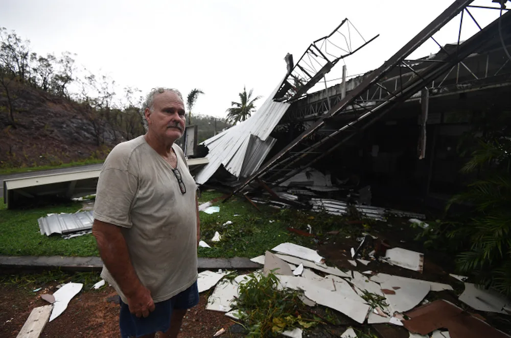 Man stands in front of a collapsed metal structure with debris scattered around after a storm.