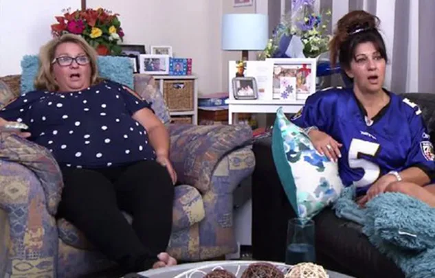 Two women sitting on couches, looking surprised, with a living room background and decorative pillows.