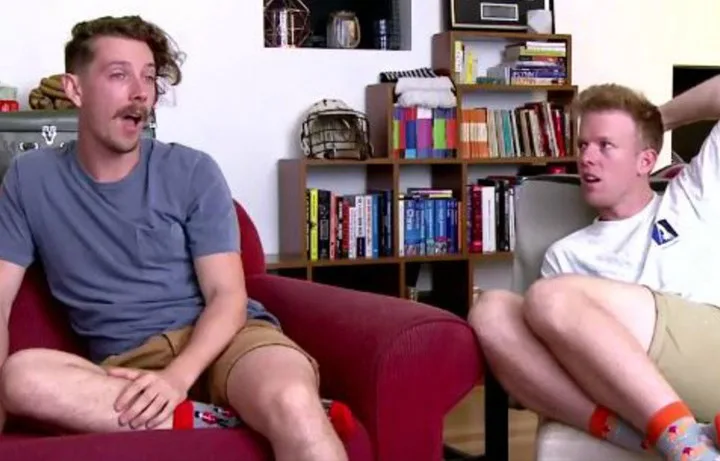 Two men sitting on chairs in a living room, talking. Shelves with books and decor are in the background.