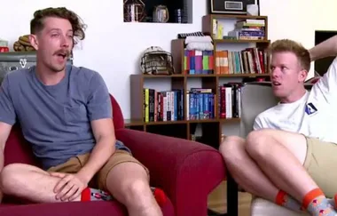 Two men sitting on chairs in a living room, talking. Shelves with books and decor are in the background.