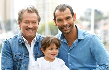 Two men smile with a young boy outdoors; all wearing blue or white shirts and surrounded by a blurred cityscape background.