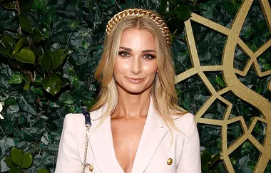 Woman in white blazer with gold headband stands in front of leafy backdrop with decorative gold design.