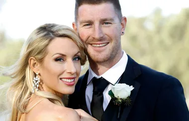 A smiling couple dressed in wedding attire, the woman wearing earrings and the man with a white boutonniere.