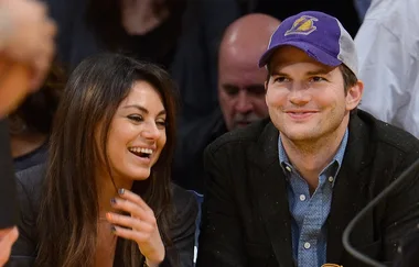Two people smiling and sitting together at a sports event, one wearing a purple and white cap.