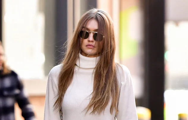 A person with long hair and sunglasses walks in an urban setting, wearing a white turtleneck sweater.