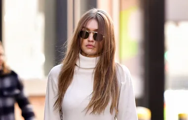 A person with long hair and sunglasses walks in an urban setting, wearing a white turtleneck sweater.