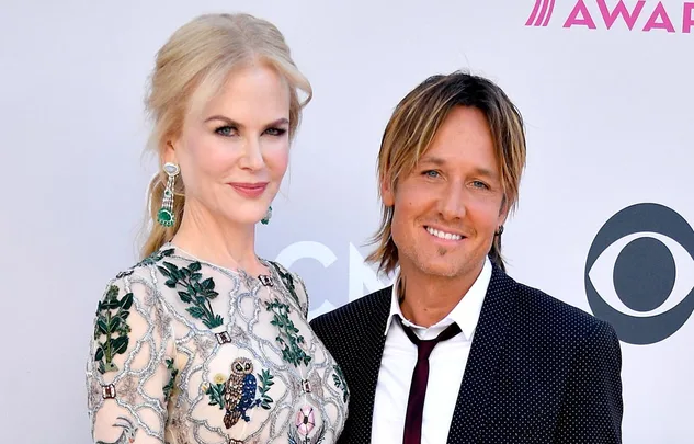 Two people pose together on a red carpet, both smiling. One wears an embroidered dress, the other a dark suit with a tie.