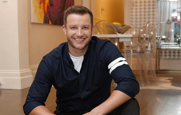 Man in a navy shirt smiling seated indoors with modern decor in the background.