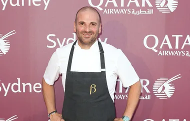 Man in a white chef coat and apron smiles against a red Qatar Airways backdrop.