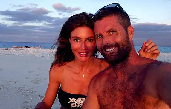 A couple taking a selfie on a sandy beach at sunset, both smiling, with the ocean and clouds in the background.