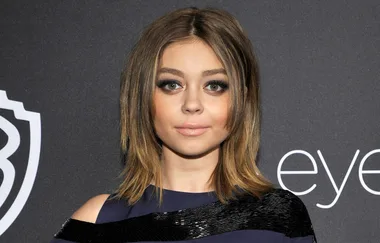 ```plaintext
Woman with shoulder-length brown hair wearing a dark dress at a formal event, standing against a branded backdrop.
```