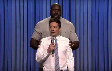 Two men on stage; one with a microphone, another standing closely behind, blue curtain backdrop.