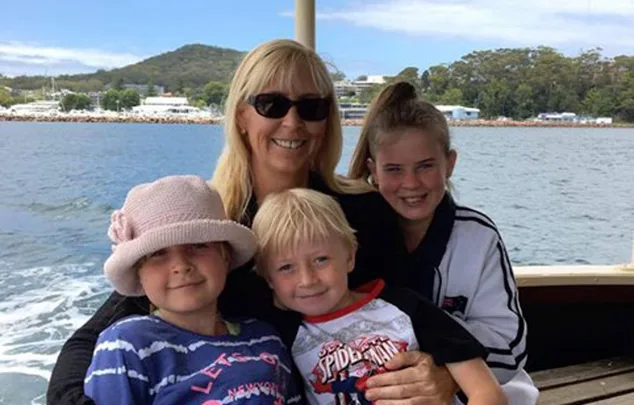 A woman and three children smile on a boat with water and trees in the background.