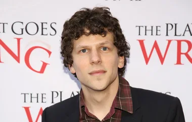 A person with curly hair, wearing a plaid shirt and blazer, stands in front of a backdrop with partial text visible.