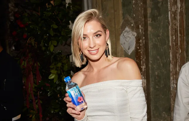 A smiling woman in an off-shoulder dress holds a bottle of FIJI Water at an event with rustic decor.
