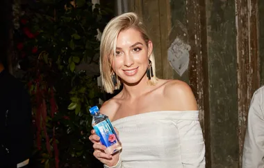 A smiling woman in an off-shoulder dress holds a bottle of FIJI Water at an event with rustic decor.