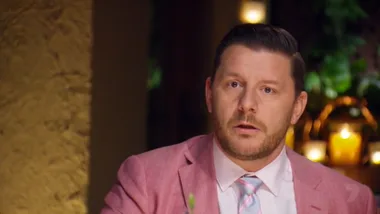 Man in a pink suit looking surprised at a dining table with candles and plants in the background.
