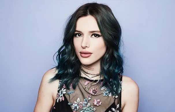 A person with blue hair wearing a floral dress and layered necklaces, set against a plain background.