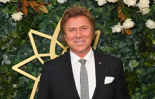 A man in a suit smiles in front of a greenery backdrop with white flowers and a geometric decoration.