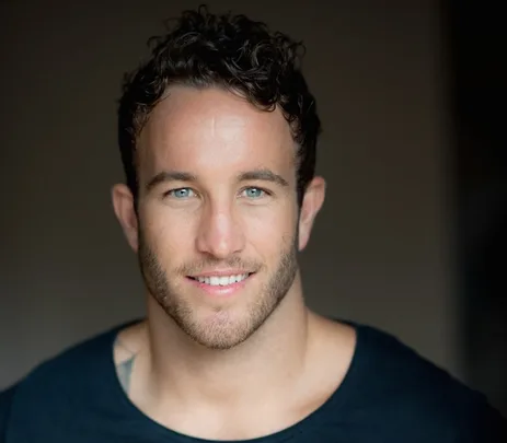 A man with short curly hair and a beard, smiling and wearing a black shirt against a dark background.