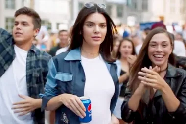 A woman holding a Pepsi can walks in a crowd, smiling.