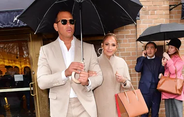 Man and woman under an umbrella, both in beige outfits, leaving a building. Woman carries a large handbag.