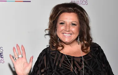 A woman in a black patterned dress smiles and waves in front of a white backdrop.