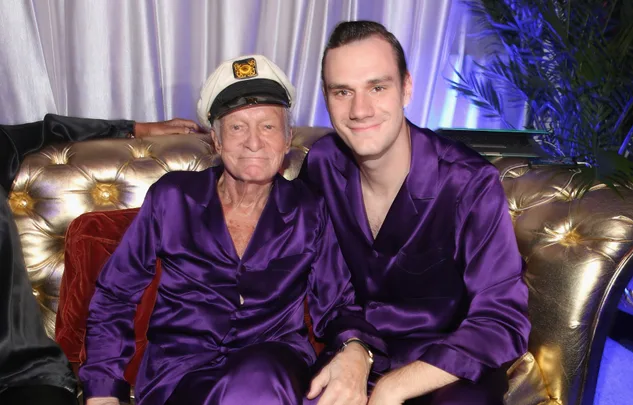 Two people in purple satin outfits sitting on a gold sofa. One wears a captain's hat, both appear content.