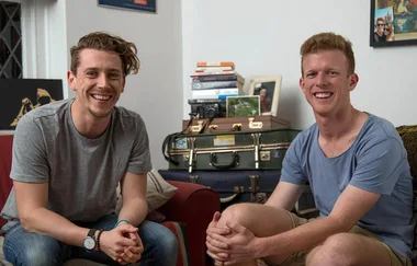 Two men smiling, seated on a sofa in a living room with books and suitcases in the background.