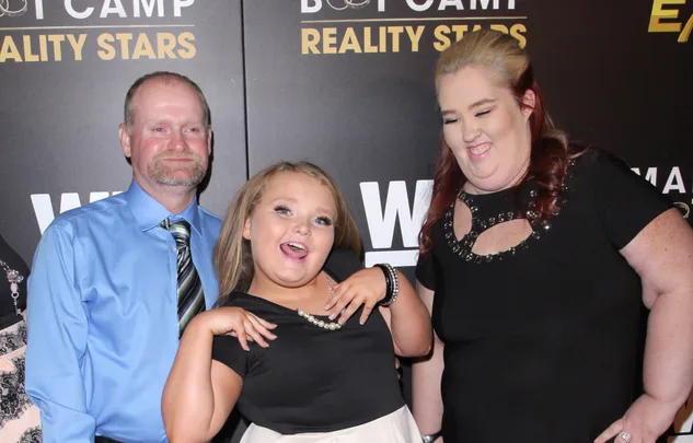 Three people posing at a red carpet event for a reality show, with logos in the background.