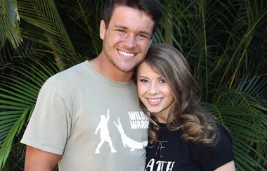 A smiling couple posing together, surrounded by lush green foliage.