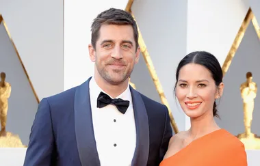Man in a tuxedo and woman in orange dress smiling on a red carpet event.