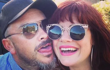 A man and woman wearing sunglasses smile outdoors, with the man playfully sticking out his tongue.