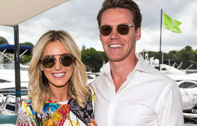 Couple smiling, wearing sunglasses, standing in front of a marina with boats in the background.