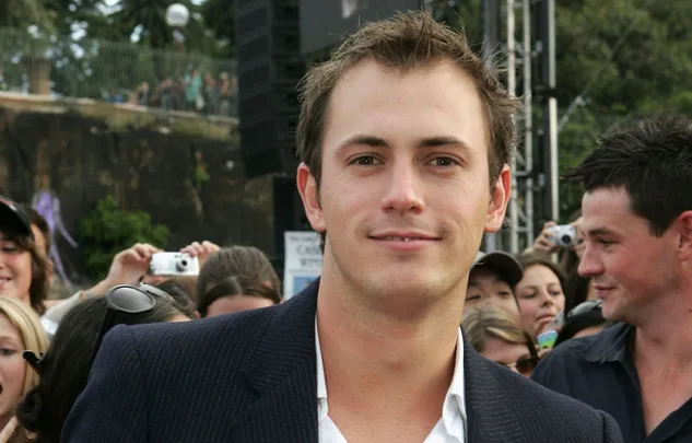 A person smiling at an outdoor event with a crowd in the background, holding cameras.