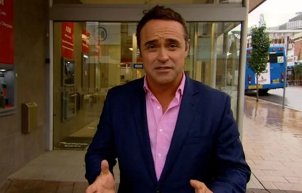 Man in blue suit and pink shirt gestures outside a building on a city street.