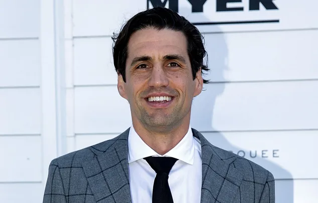 A man in a gray checkered suit and black tie, smiling against a white background.