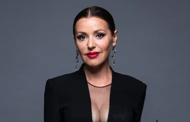 A woman with dark hair and red lipstick, wearing drop earrings and a black outfit against a gray background.