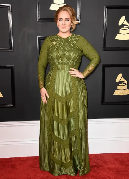 Person in an elegant green gown on a red carpet at a formal event, with framed Grammy symbols in the background.