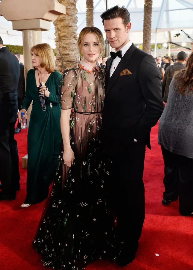 A man in a tuxedo and a woman in a floral dress pose together on a red carpet, surrounded by other guests.