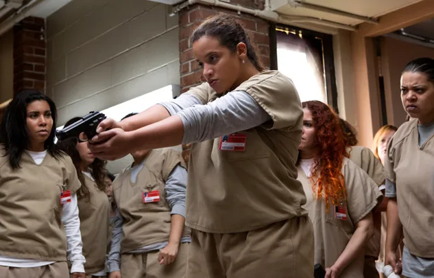 Prison inmate in beige uniform aims a gun while others watch tensely in a correctional facility setting.