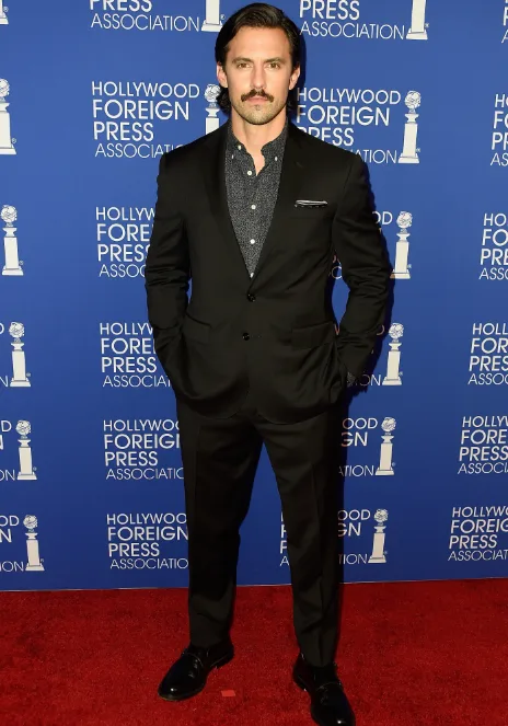 A man in a black suit with a dotted shirt and mustache poses on the red carpet at a Hollywood Foreign Press event.