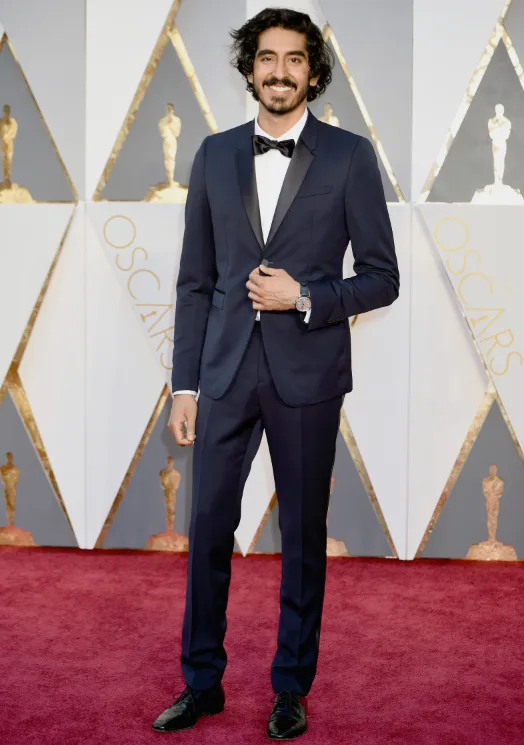 Man in a dark suit with bowtie on a red carpet at an award event with Oscar statues in the background.