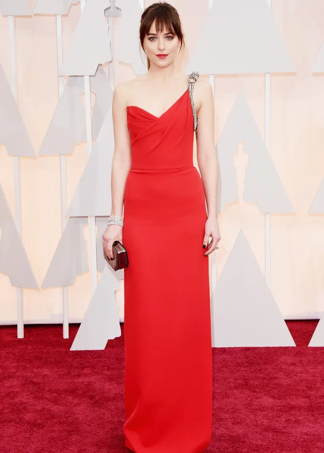 A woman in a red one-shoulder gown stands on a red carpet holding a clutch, with a geometric backdrop.