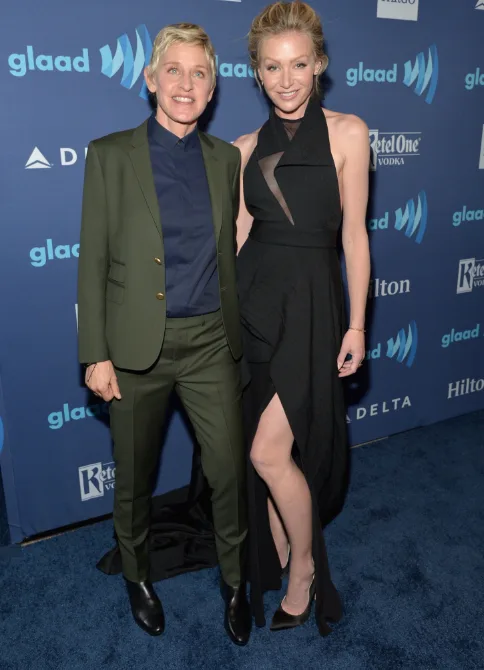 Two people smiling and posing on a blue carpet at the GLAAD Awards, one in a green suit and the other in a black dress.