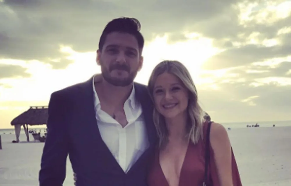 A man in a suit and a woman in a red dress smile on a beach at sunset.